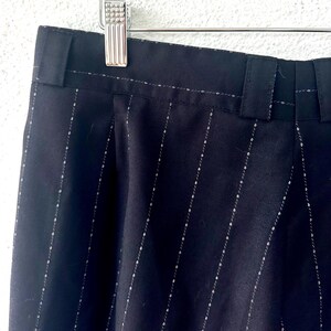 Peut inclure: Pantalon noir avec un motif discret de fines rayures blanches. Le v&ecirc;tement comprend une ceinture avec des passants int&eacute;gr&eacute;s et est suspendu &agrave; un cintre en m&eacute;tal argent&eacute;. Le tissu pr&eacute;sente une texture lisse avec des lignes verticales.