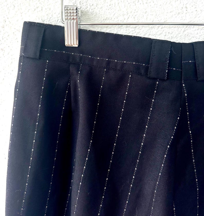 Peut inclure: Pantalon noir avec un motif discret de fines rayures blanches. Le v&ecirc;tement comprend une ceinture avec des passants int&eacute;gr&eacute;s et est suspendu &agrave; un cintre en m&eacute;tal argent&eacute;. Le tissu pr&eacute;sente une texture lisse avec des lignes verticales.