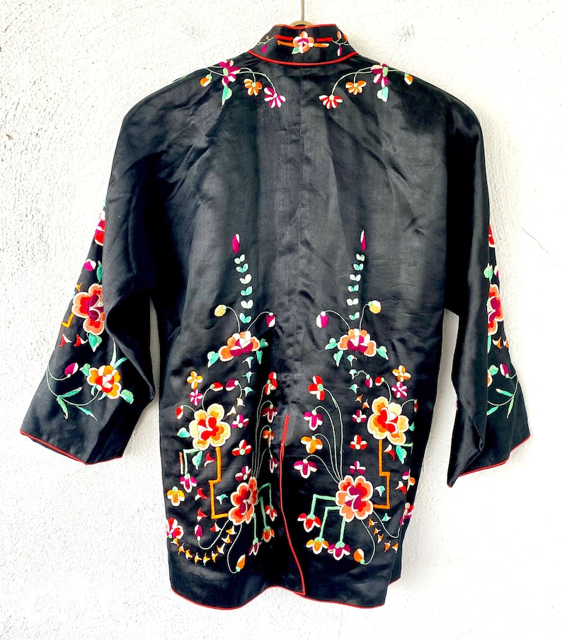 Puede incluir: Una chaqueta de seda negra con cuello mandar&iacute;n y bordado floral intrincado en tonos de rojo, rosa, verde y amarillo.