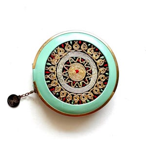 May include: A round, mint green compact mirror with a gold trim. The mirror has a decorative design in the center, featuring a gold and silver mandala with red and green accents.