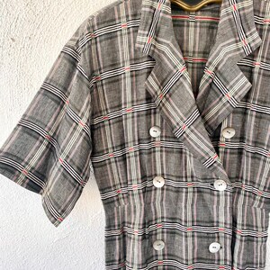 May include: A short-sleeved, double-breasted dress featuring a grey, black, white, and red plaid pattern. The garment has a notched lapel collar and six round, light-colored buttons arranged in two vertical rows down the front bodice.