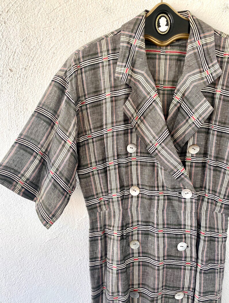 May include: A short-sleeved, double-breasted dress featuring a grey, black, white, and red plaid pattern. The garment has a notched lapel collar and six round, light-colored buttons arranged in two vertical rows down the front bodice.
