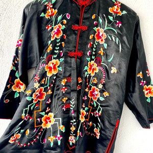 Puede incluir: Una bata de seda negra con ribetes rojos y bordados florales. La bata tiene un cuello mandar&iacute;n y un cierre frontal con botones decorativos.