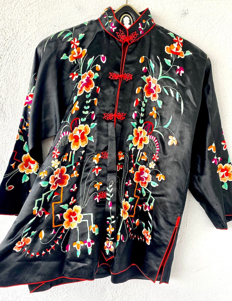 Puede incluir: Una bata de seda negra con ribetes rojos y bordados florales. La bata tiene un cuello mandar&iacute;n y un cierre frontal con botones decorativos.