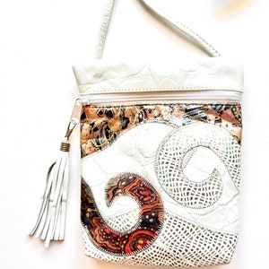 May include: White leather crossbody bag with a patchwork design featuring swirls and geometric patterns in shades of brown, orange, and white. The bag has a zipper closure and a white tassel.