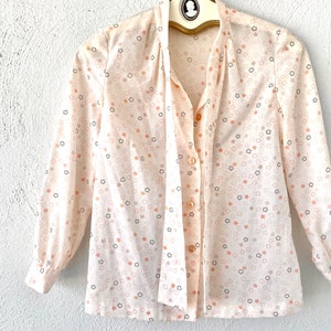 May include: A cream-colored blouse with a star pattern in peach and black. The long-sleeved shirt has a tie-neck design and is buttoned down the front. The blouse is hanging on a vintage hanger.