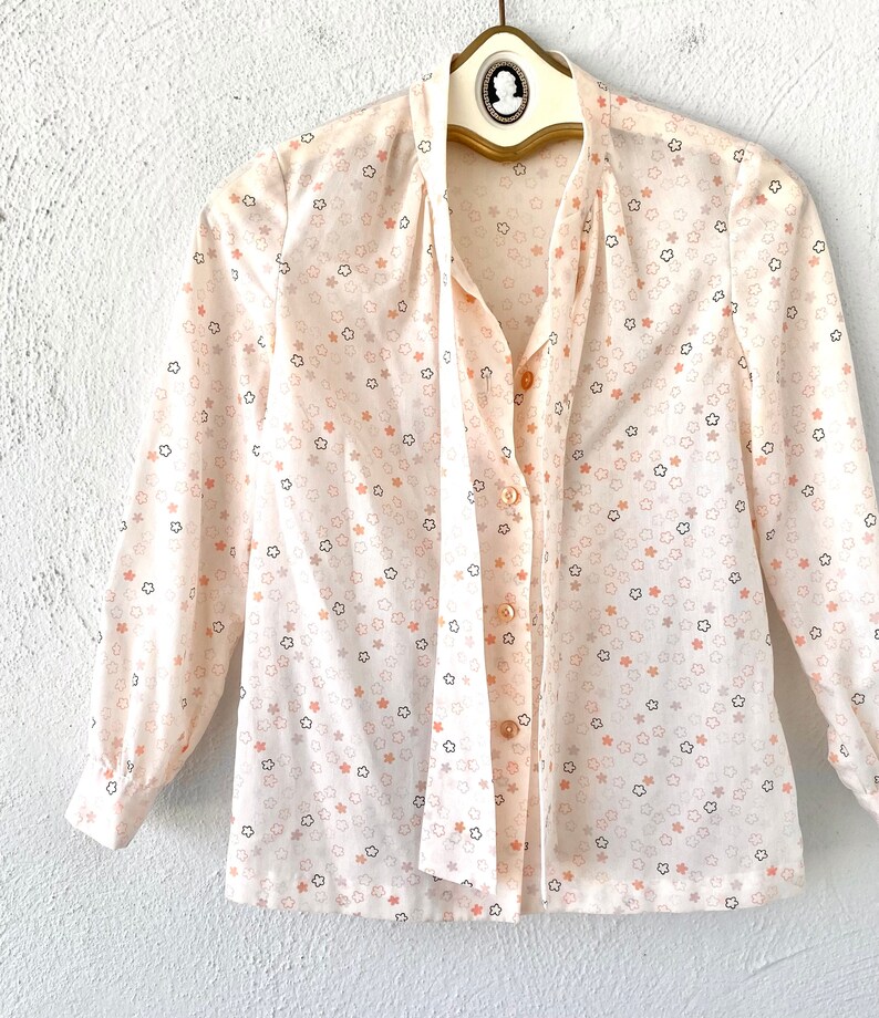 May include: A cream-colored blouse with a star pattern in peach and black. The long-sleeved shirt has a tie-neck design and is buttoned down the front. The blouse is hanging on a vintage hanger.
