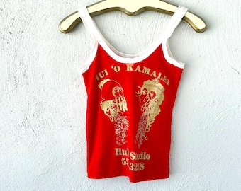 May include: Red tank top with white trim and straps, featuring gold text and graphics. The text reads "Hui 'O Kamalei" and "Hul Studio 53218". The tank top is hanging on a gold-colored hanger.