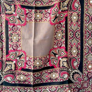 May include: A square scarf with an intricate paisley design. The scarf features a central tan panel surrounded by a border of black, pink, gold, and gray paisley patterns. The edges are finished with a fringe.