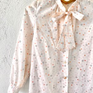 May include: A cream-colored blouse with a floral pattern of peach, gray, and black flowers. The long-sleeved shirt features a bow tie at the neckline and is buttoned down the front. The blouse is hanging on a gold hanger.