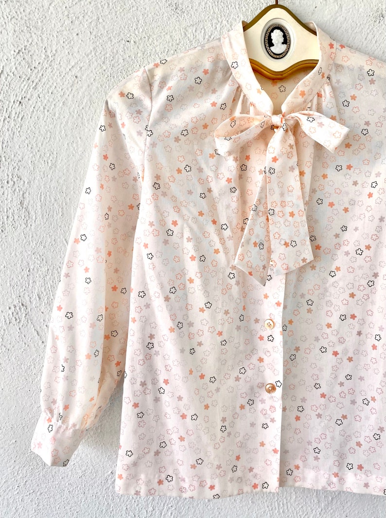 May include: A cream-colored blouse with a floral pattern of peach, gray, and black flowers. The long-sleeved shirt features a bow tie at the neckline and is buttoned down the front. The blouse is hanging on a gold hanger.