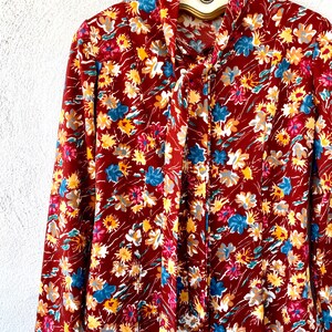 Pode incluir: Uma blusa de manga comprida com um la&ccedil;o no pesco&ccedil;o. A blusa &eacute; feita de um tecido marrom com uma estampa floral em tons de vermelho, amarelo, azul e branco.
