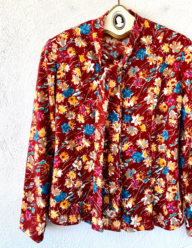 Pode incluir: Uma blusa de manga comprida com um la&ccedil;o no pesco&ccedil;o. A blusa &eacute; feita de um tecido marrom com uma estampa floral em tons de vermelho, amarelo, azul e branco.