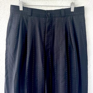 Peut inclure: Pantalon noir avec un motif discret de fines rayures verticales ponctu&eacute;es de petits points blancs. Le pantalon pr&eacute;sente une ceinture avec des passants, une fermeture &agrave; bouton centrale et des plis verticaux partant de la taille.
