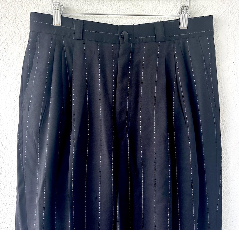 Peut inclure: Pantalon noir avec un motif discret de fines rayures verticales ponctu&eacute;es de petits points blancs. Le pantalon pr&eacute;sente une ceinture avec des passants, une fermeture &agrave; bouton centrale et des plis verticaux partant de la taille.