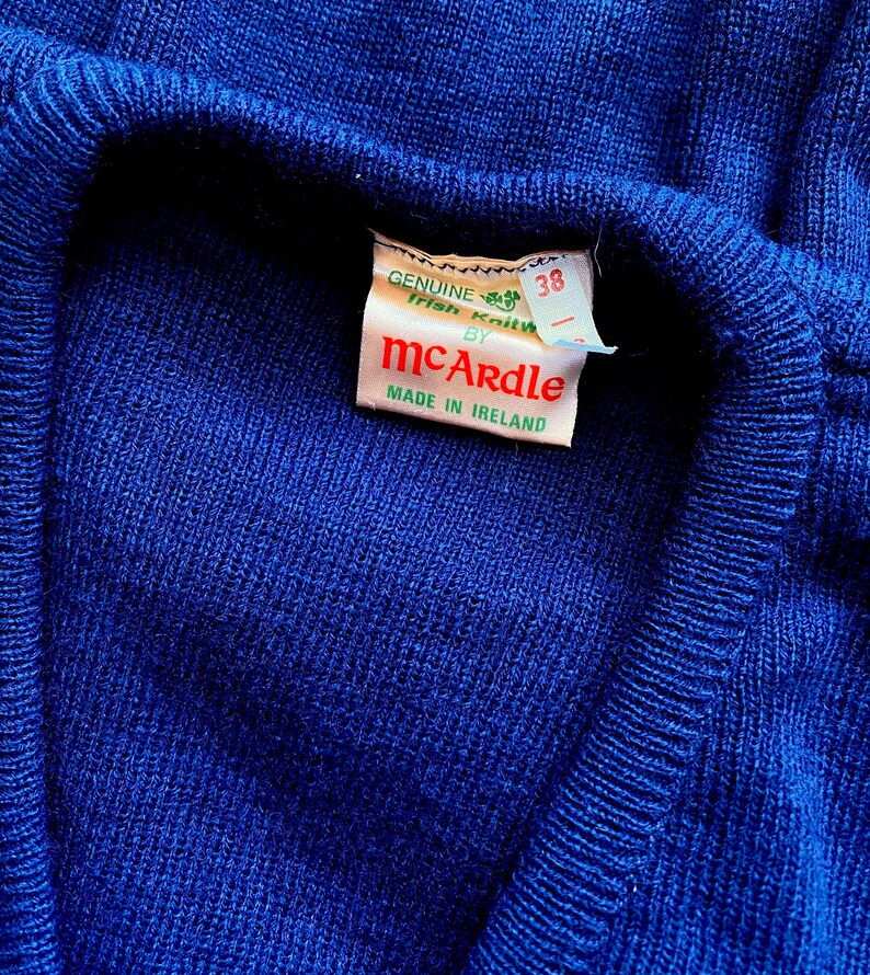 Może przedstawiać: Niebieski sweter z białym znaczkiem z napisem "GENUINE Irish Knitwear by McArdle MADE IN IRELAND" i metką rozmiaru z napisem "38".