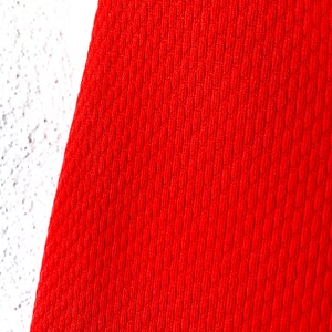 May include: Close-up of a vibrant red fabric with a textured, ribbed pattern. The fabric is set against a white, textured surface. The red material appears to be a knit or woven textile.