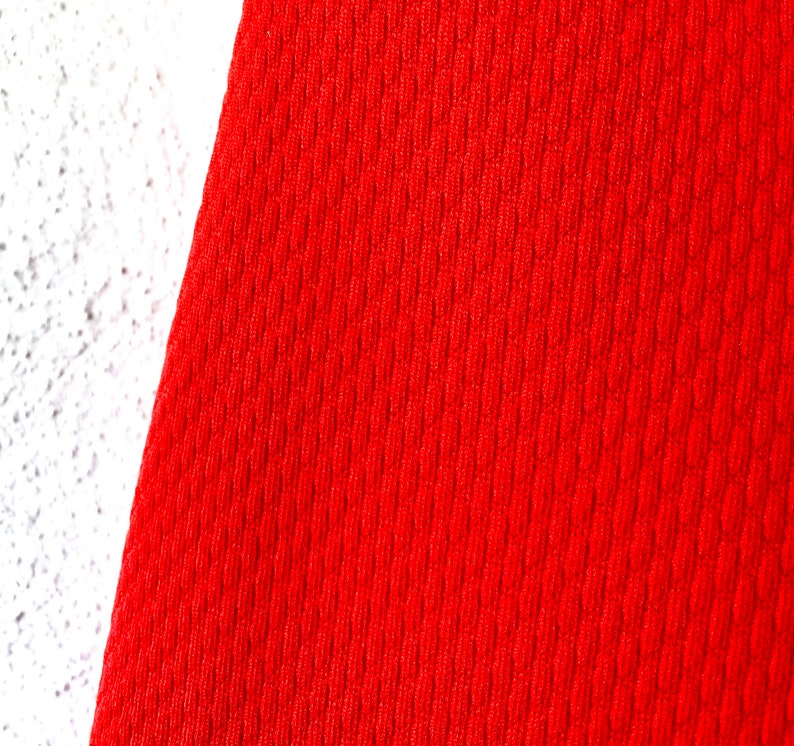 May include: Close-up of a vibrant red fabric with a textured, ribbed pattern. The fabric is set against a white, textured surface. The red material appears to be a knit or woven textile.