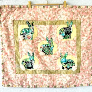 May include: A pink and green patchwork quilt with a floral pattern and five bunny rabbits in the center. The bunnies are made of different fabrics and have a green and brown floral pattern.