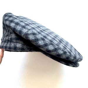 May include: A grey and black plaid newsboy cap. The cap is made of a soft, textured fabric and features a classic, rounded shape with a small brim. The pattern is a subtle, checkered design.