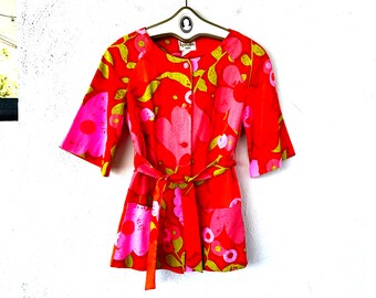 Pode incluir: Uma jaqueta de manga curta com estampa floral vermelha e rosa com cinto de laço. A jaqueta tem fecho de botão e dois bolsos.