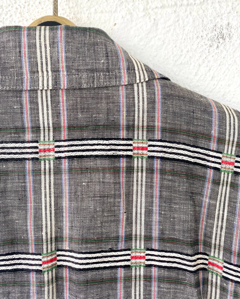 May include: A charcoal gray shirt featuring a textured weave with vertical pinstripes in white, blue, and red. The fabric is overlaid with horizontal bands of white and black stripes, accented by small squares containing red and green thread details.
