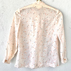 May include: A cream-colored blouse with a star pattern in shades of peach, gray, and black. The long-sleeved shirt has a button-down front and cuffs, and is hanging on a gold-colored hanger.