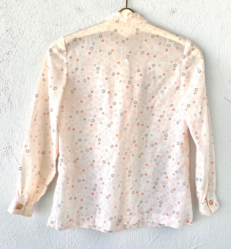 May include: A cream-colored blouse with a star pattern in shades of peach, gray, and black. The long-sleeved shirt has a button-down front and cuffs, and is hanging on a gold-colored hanger.