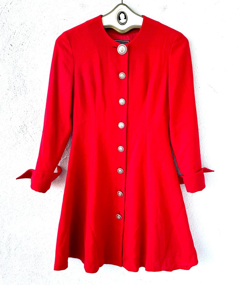 Puede incluir: Un abrigo rojo brillante con mangas largas y falda acampanada. El abrigo presenta una fila de botones plateados grandes y redondos y pu&ntilde;os decorativos. Cuelga de una percha vintage.