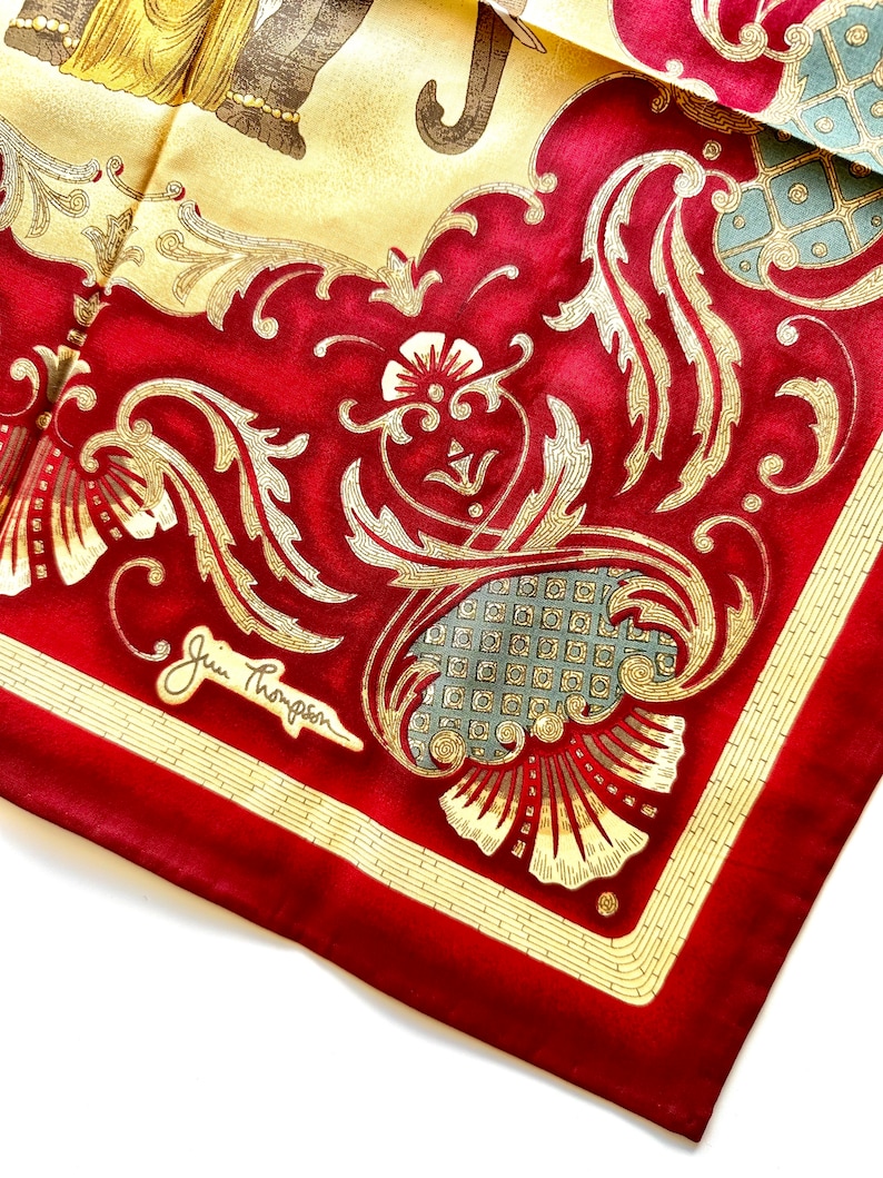 May include: A silk scarf with a vibrant red border and a central design featuring gold filigree and floral patterns. The scarf has a cream-colored background with an elephant illustration. The signature "Jim Thompson" is visible.