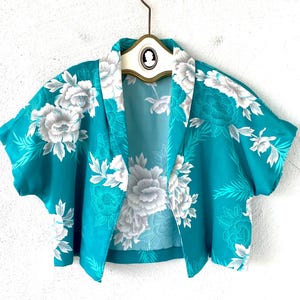 May include: A teal blue cropped jacket with a white floral print. The jacket has a collar and short sleeves.