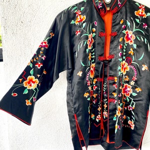 Puede incluir: Una chaqueta de seda negra con ribetes rojos y bordados florales de colores. La chaqueta tiene un cuello mandar&iacute;n y un cierre frontal con botones decorativos.