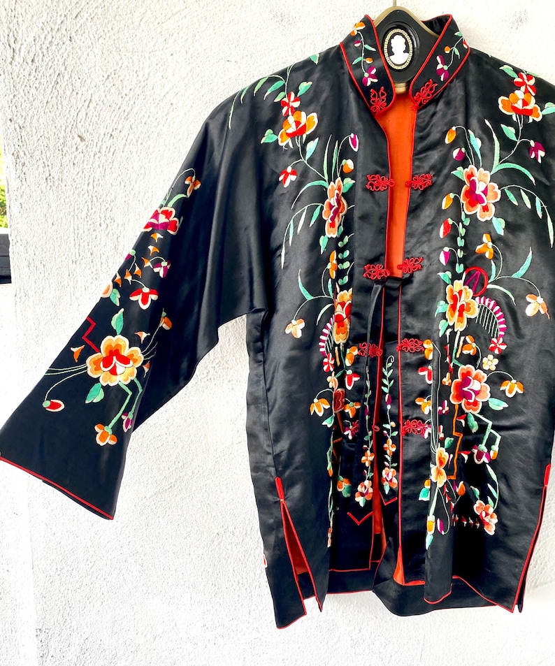 Puede incluir: Una chaqueta de seda negra con ribetes rojos y bordados florales de colores. La chaqueta tiene un cuello mandar&iacute;n y un cierre frontal con botones decorativos.