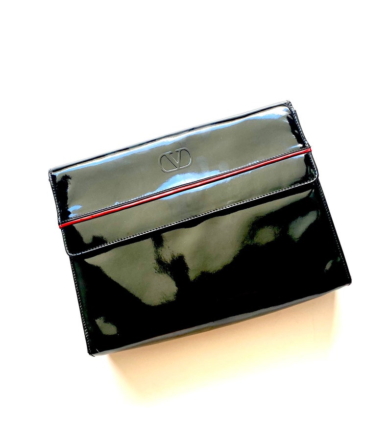 Op de afbeelding: Een zwarte clutch van gelakt leer met een rode bies en een reli&euml;f "VD" logo.