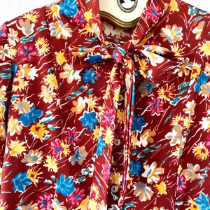 Pode incluir: Uma blusa marrom de manga comprida com estampa floral com flores azuis, amarelas e rosas. A blusa tem um la&ccedil;o no pesco&ccedil;o e fechamentos de bot&atilde;o.