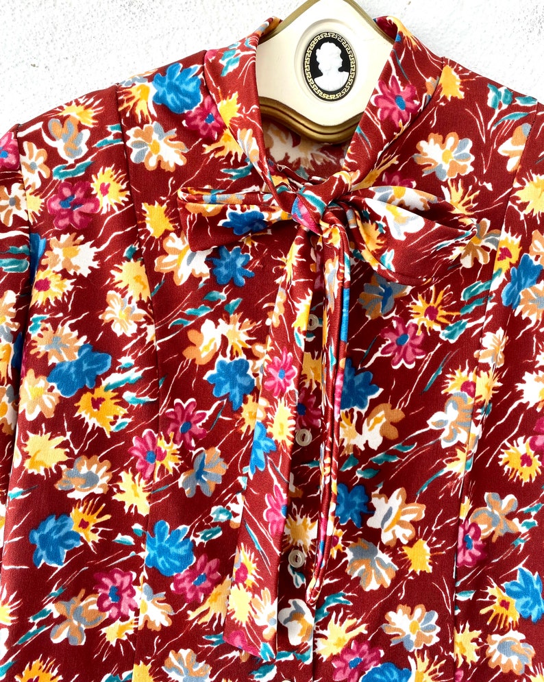 Pode incluir: Uma blusa marrom de manga comprida com estampa floral com flores azuis, amarelas e rosas. A blusa tem um la&ccedil;o no pesco&ccedil;o e fechamentos de bot&atilde;o.