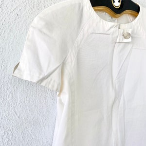 May include: White short-sleeved button-up shirt with a round neckline and a single button closure. The shirt has a slightly fitted silhouette and a relaxed fit.
