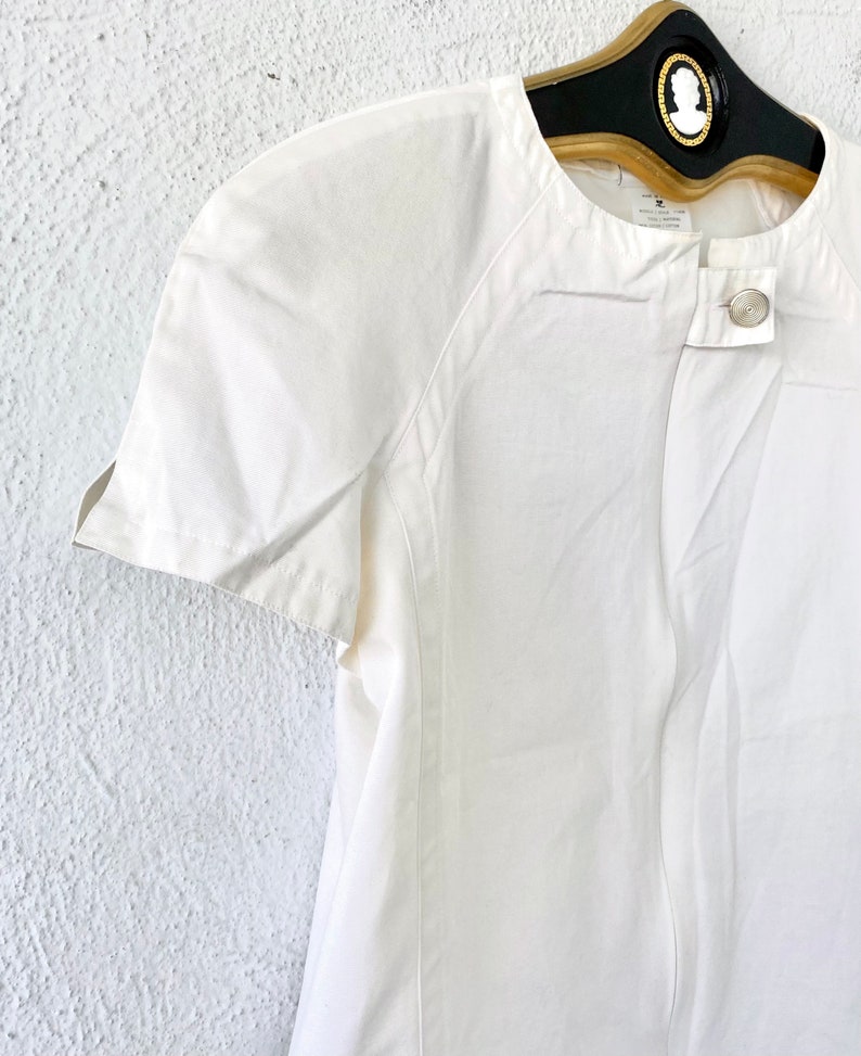 May include: White short-sleeved button-up shirt with a round neckline and a single button closure. The shirt has a slightly fitted silhouette and a relaxed fit.