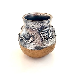 Op de afbeelding: Een kleine, ronde, bruine kleipot met een zilveren glazuur. De pot heeft een ontwerp met een gezicht met een hoofdtooi en een slang aan de zijkant.
