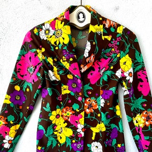 May include: A brown vintage jacket with a floral print featuring pink, yellow, white, orange, and purple flowers. The jacket has a long sleeve and a button closure.