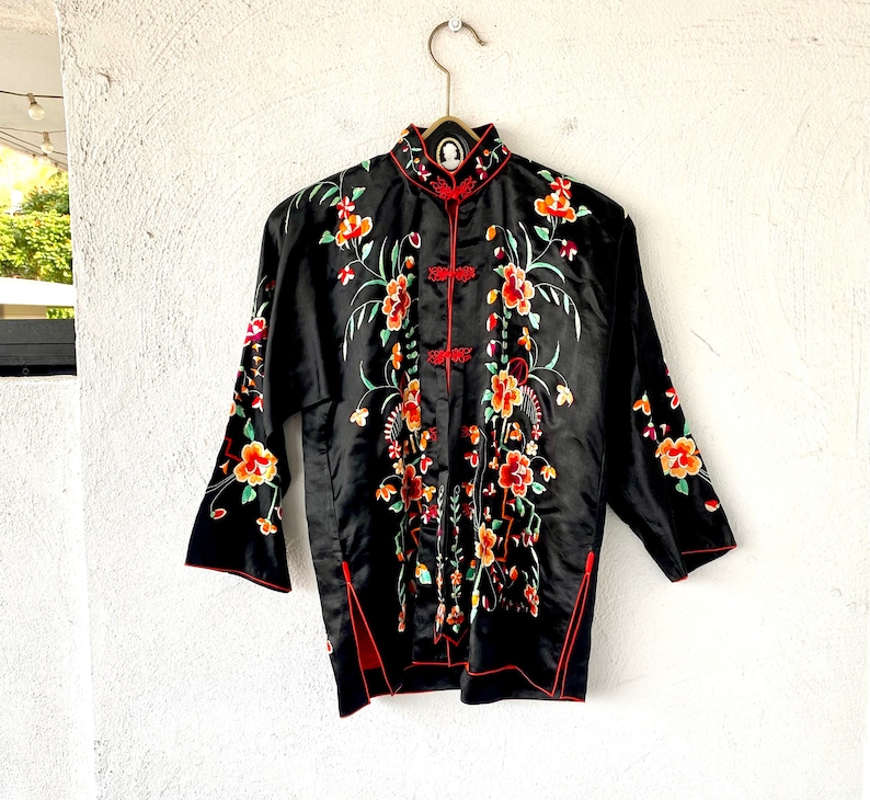 Puede incluir: Chaqueta de seda negra con bordado floral rojo y verde. La chaqueta tiene un cuello mandar&iacute;n y cierre de bot&oacute;n.