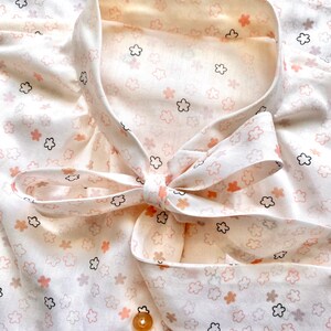 May include: A cream-colored blouse with a delicate floral pattern in shades of peach, black, and brown. The blouse features a bow tie collar and a single gold button. The fabric has a soft, flowing appearance.