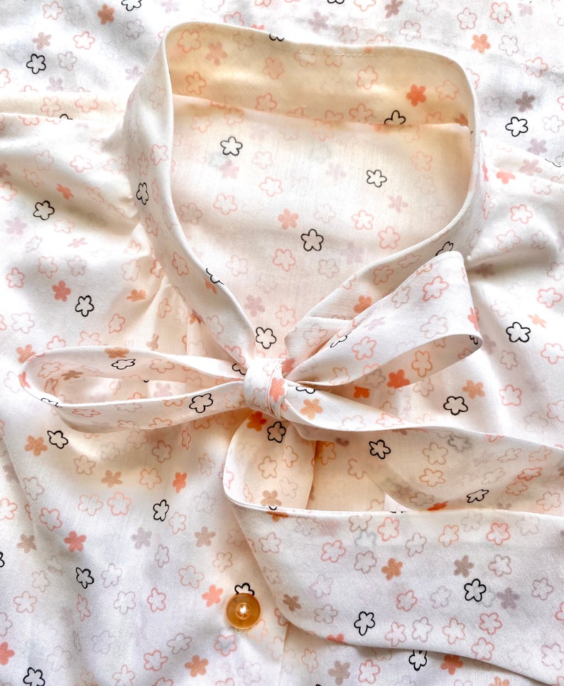 May include: A cream-colored blouse with a delicate floral pattern in shades of peach, black, and brown. The blouse features a bow tie collar and a single gold button. The fabric has a soft, flowing appearance.