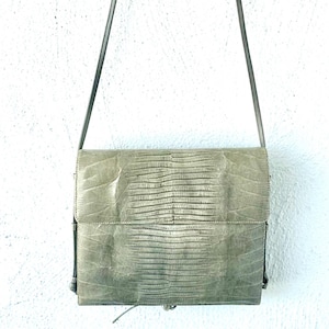 May include: A sage green rectangular shoulder bag featuring a textured lizard skin pattern on the front flap and body. The bag includes a thin, matching shoulder strap attached to the top sides, with a structured silhouette and a clean finish.