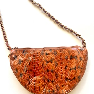 May include: A crescent-shaped handbag featuring a textured, orange and brown snakeskin pattern. The bag includes a top zip closure and a long shoulder strap made of a gold-tone metal chain interwoven with a thin brown leather strip.