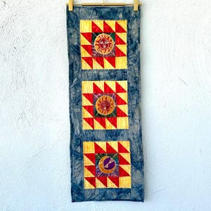 May include: A decorative wall hanging featuring three panels with sun and moon designs. Each panel has a yellow and red geometric pattern with a central sun or moon image. The background is a tie-dye blue fabric.