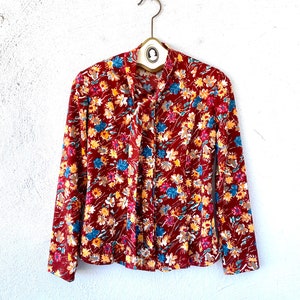 Pode incluir: Uma blusa de manga comprida vermelha com estampa floral com flores amarelas, azuis e brancas. A blusa tem um la&ccedil;o no pesco&ccedil;o.