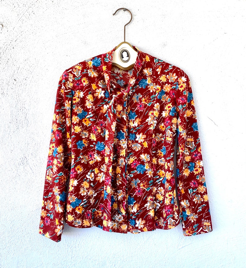 Pode incluir: Uma blusa de manga comprida vermelha com estampa floral com flores amarelas, azuis e brancas. A blusa tem um la&ccedil;o no pesco&ccedil;o.