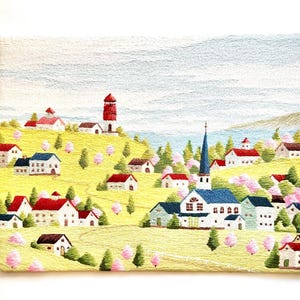May include: Embroidered wall art depicting a quaint village with colorful houses, a church with a blue steeple, and a red lighthouse on a hill. The scene is set against a backdrop of a blue sky with white clouds and a body of water.