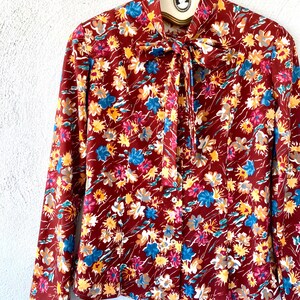 Pode incluir: Uma blusa de manga comprida com um la&ccedil;o no pesco&ccedil;o. A blusa &eacute; feita de um tecido marrom com uma estampa floral em tons de vermelho, azul, amarelo e branco.